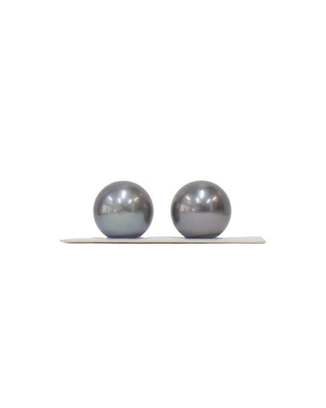 Two smooth, dark grey, spherical pearls are resting side-by-side on a thin, white surface against a plain white background.