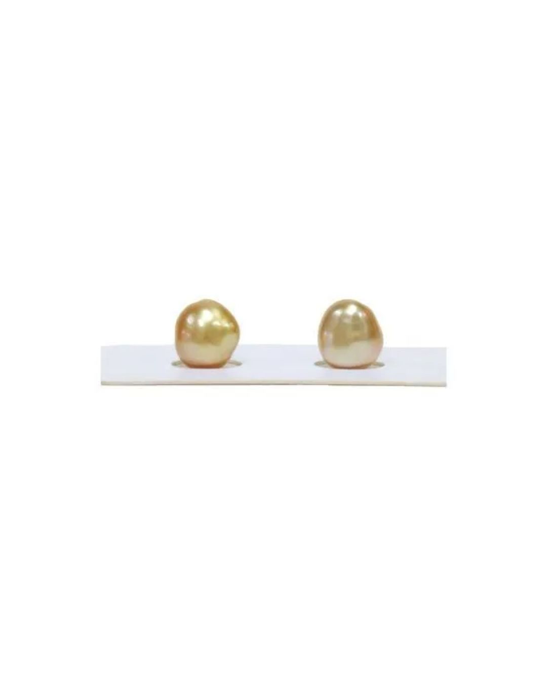 Two golden-hued, irregularly shaped baroque pearls resting side by side in indentations on a white display card.