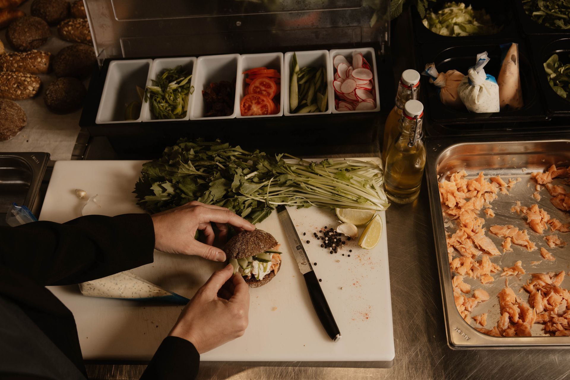 Handen die een broodje klaarmaken in de keuken, met gesneden groenten, kruiden en andere ingrediënten in de buurt.