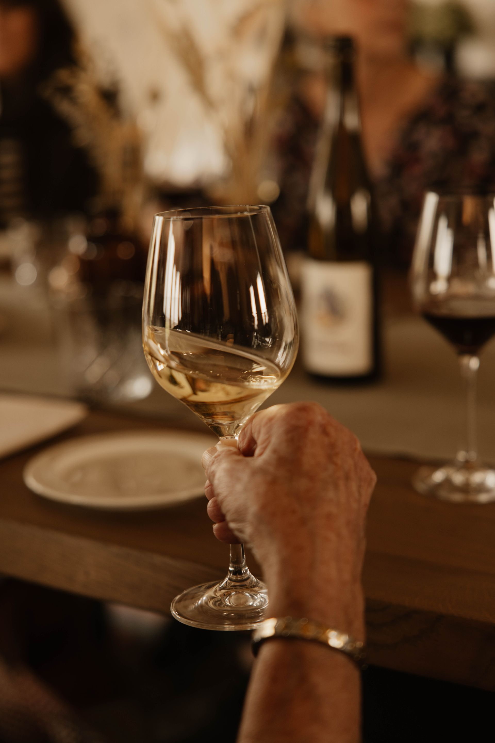 Een hand houdt een wijnglas vast aan een eettafel bij kaarslicht, met wazige glazen en flessen op de achtergrond.