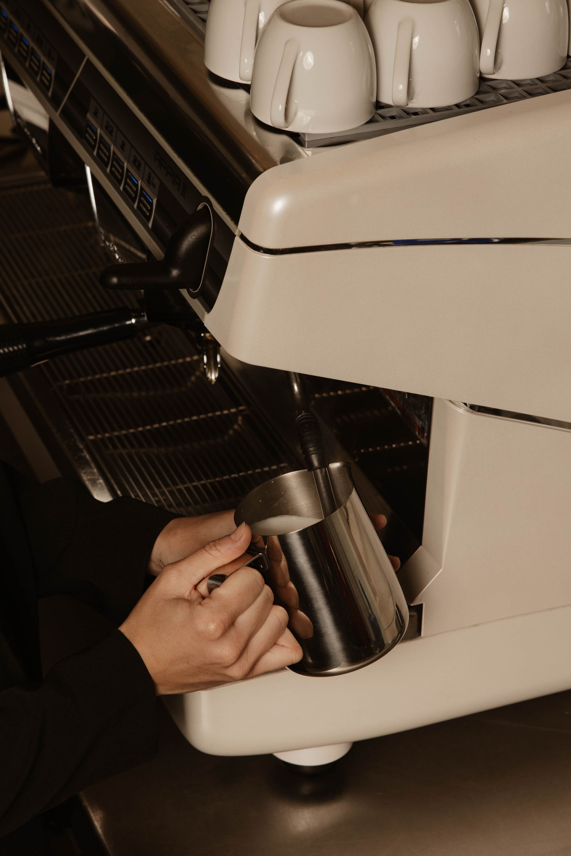 Een hand plaatst een metalen melkkan onder de stoompijp van een espressomachine.