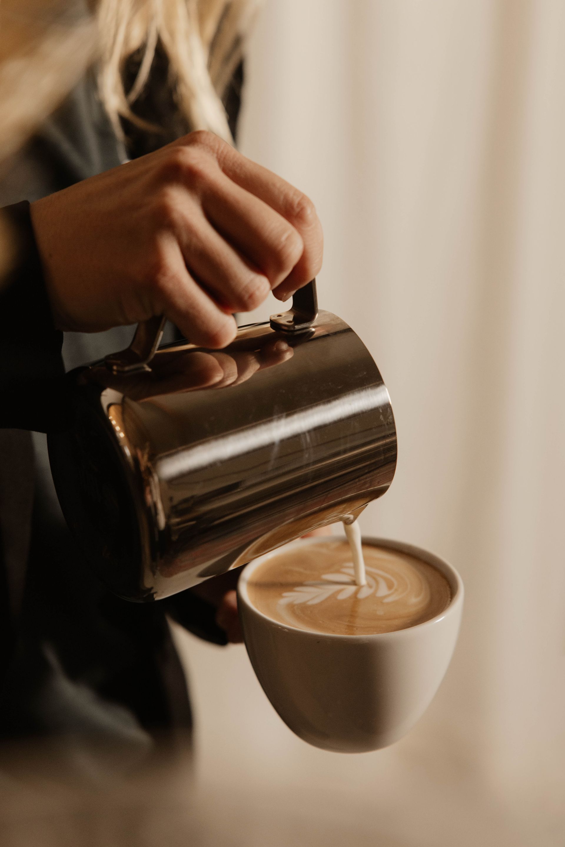 Koffie wordt met de hand vanuit een metalen kan in een wit kopje geschonken, waarbij de latte art zichtbaar is.