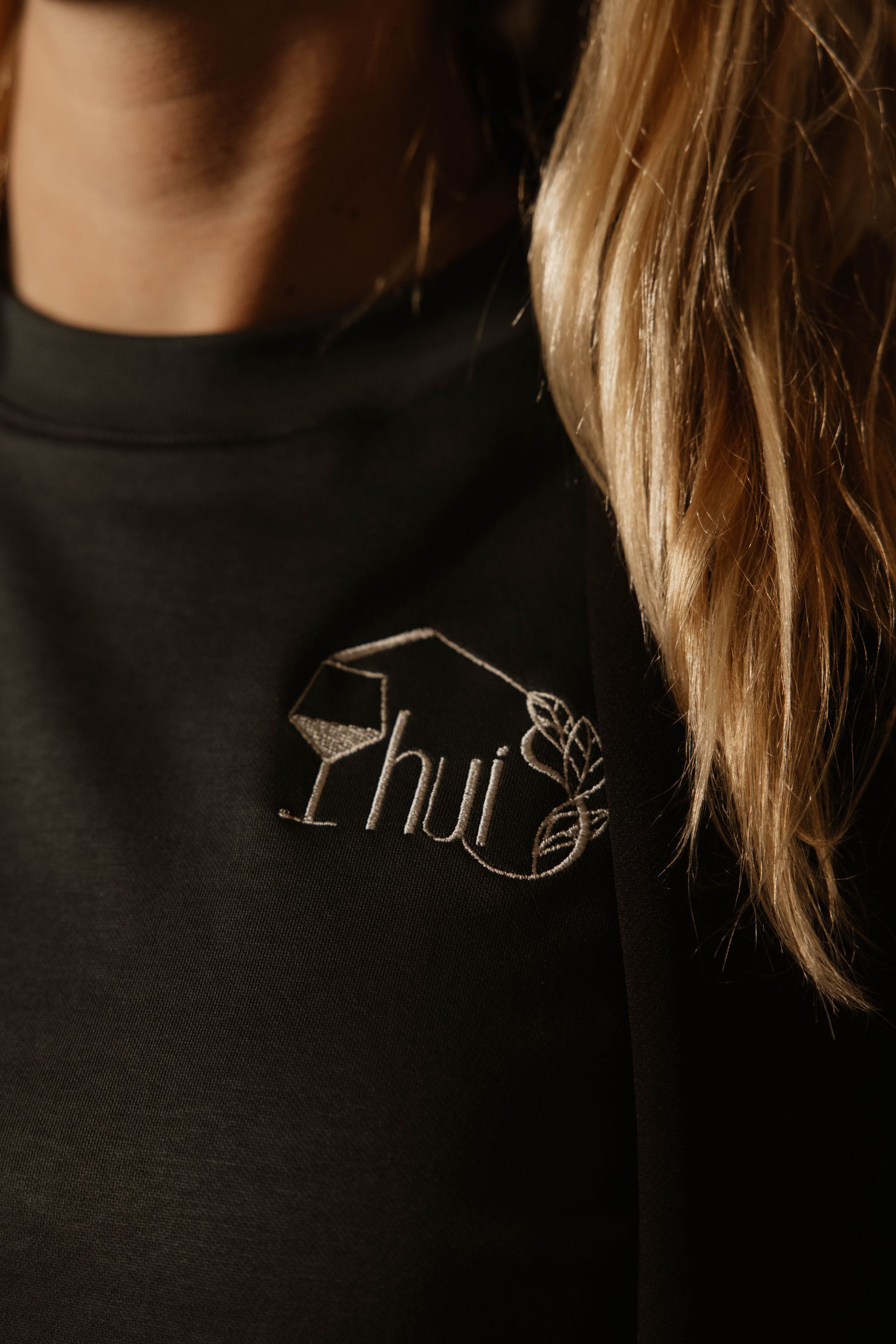 Close-up van een donkergroen shirt met een klein logo en een blonde haarlok aan de rechterkant.