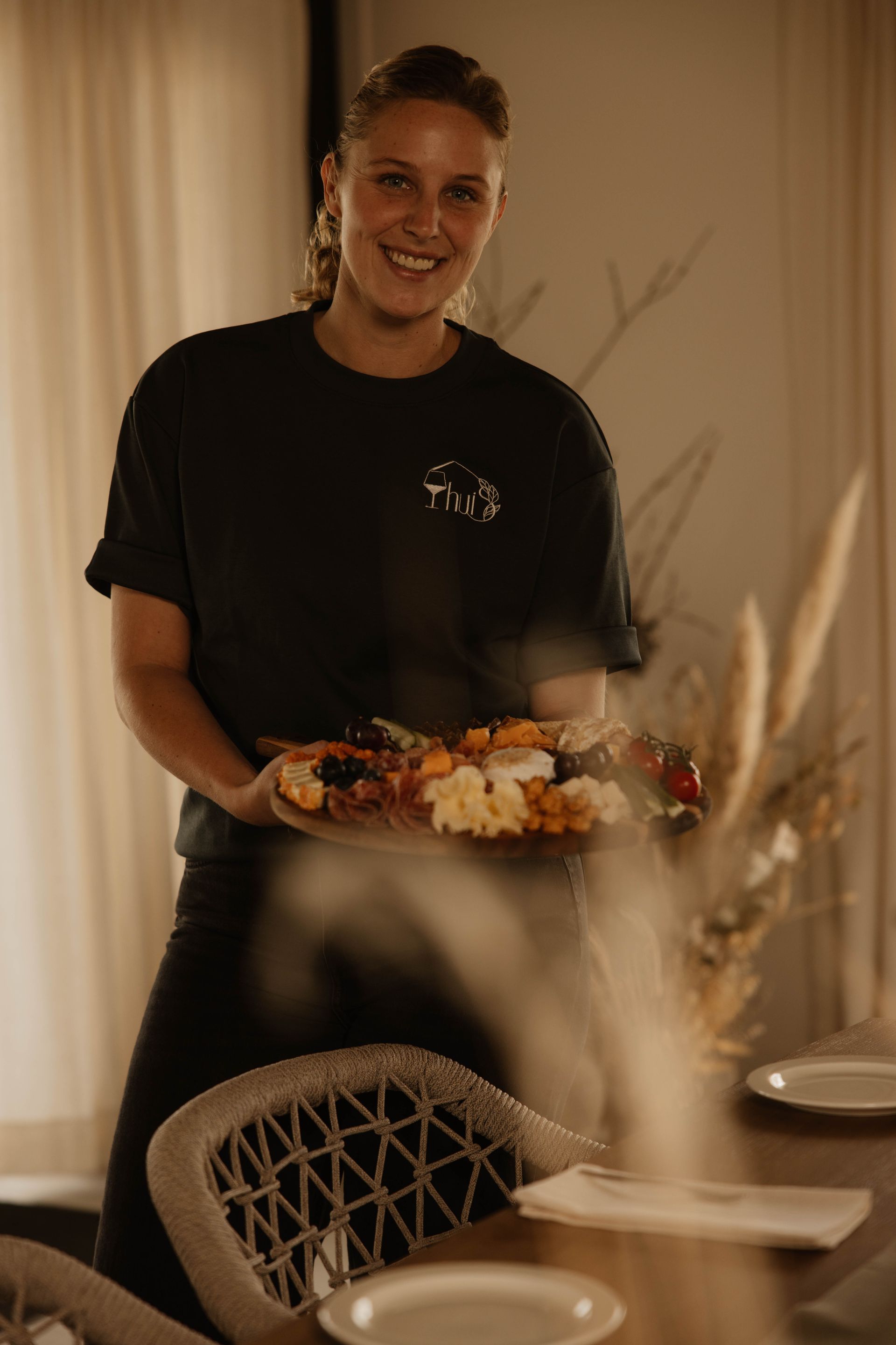 Yara lacht en houdt een dienblad met eten vast in een sfeervol verlichte eetkamer.