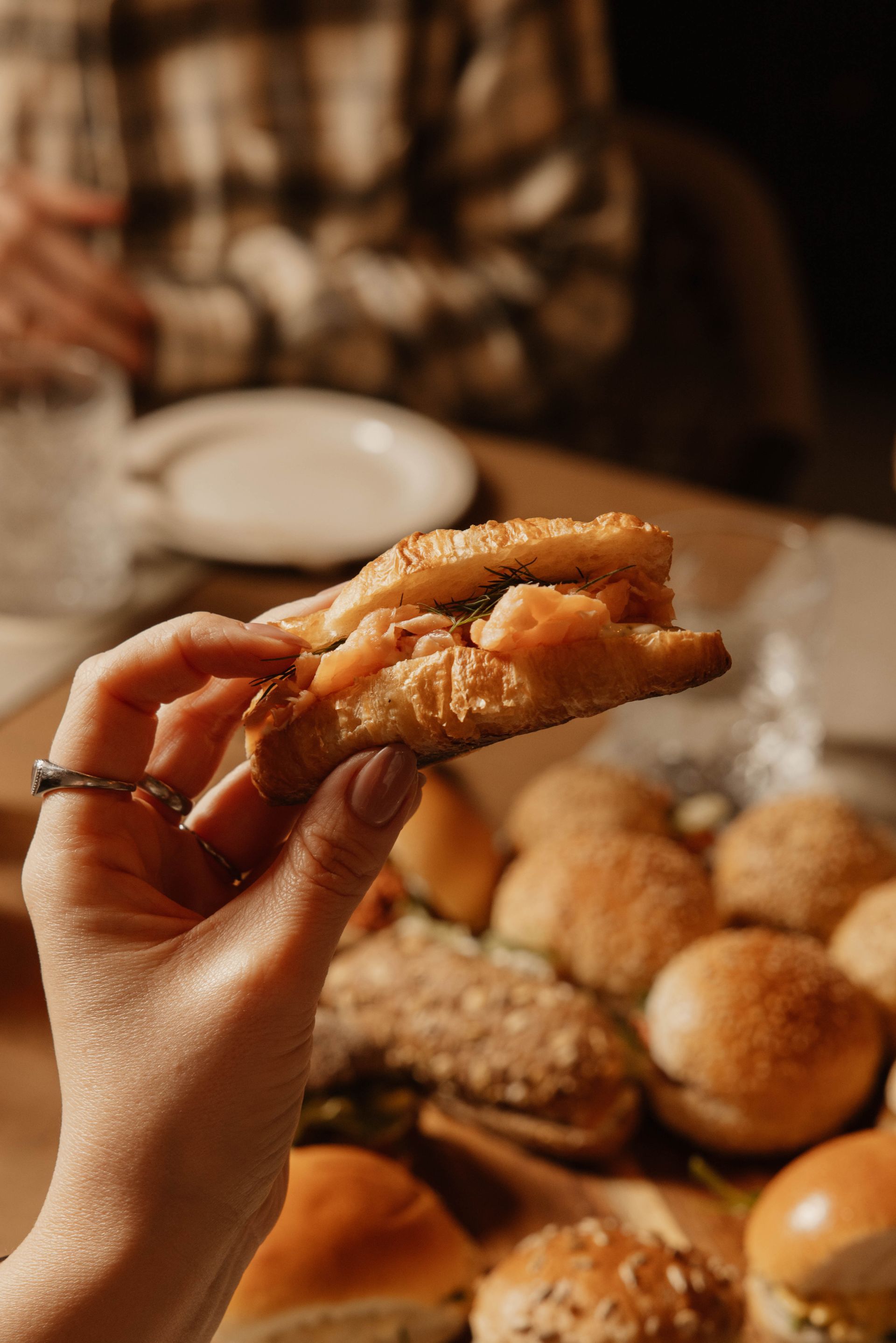 Hand die een croissantje met zalmforel vasthoudt boven een tafel vol diverse broodjes in warm licht.
