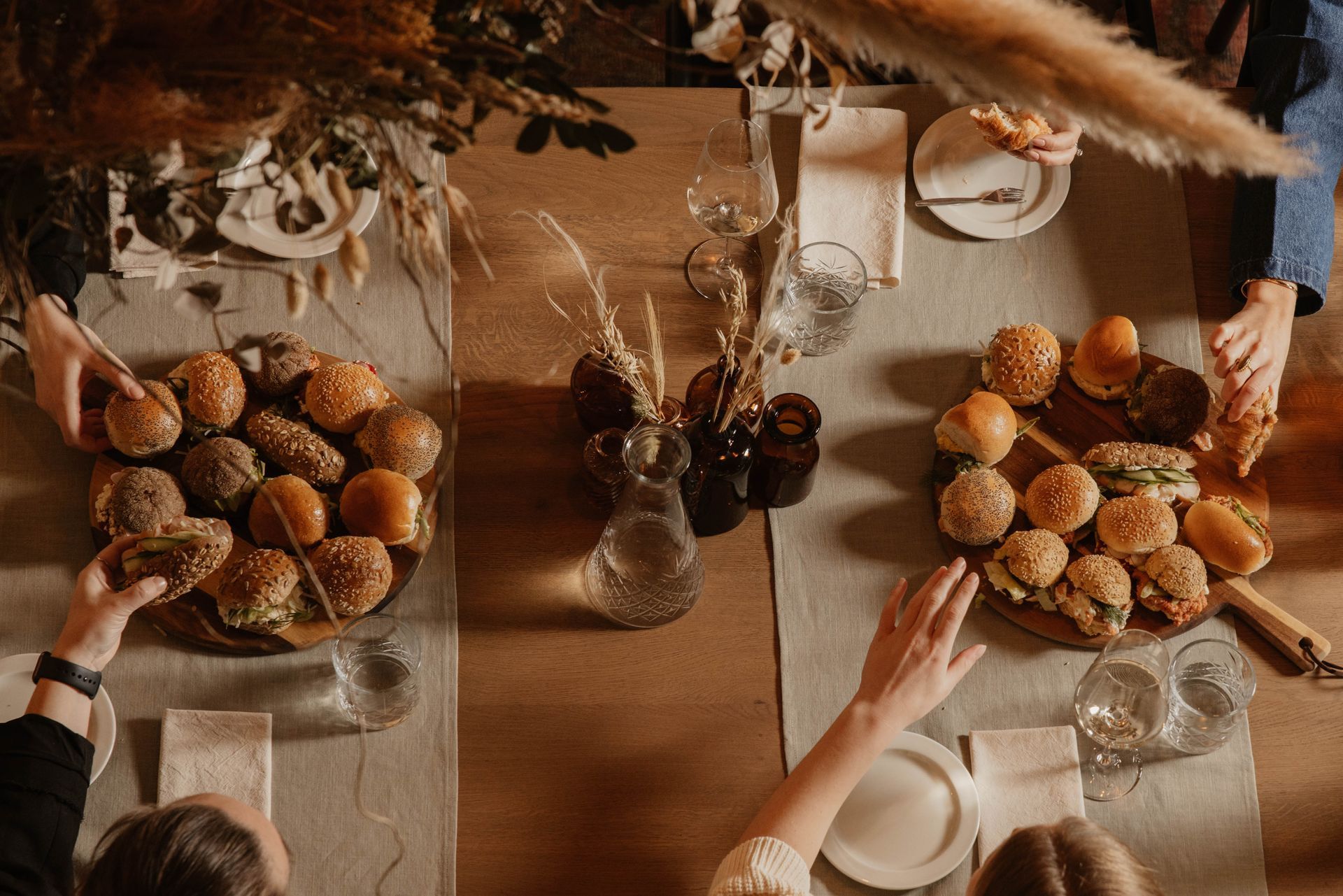 Twee mensen pakken een broodje van een rustieke tafel met broodplanken, borden, kaarsen en gedroogde bloemen als decoratie.