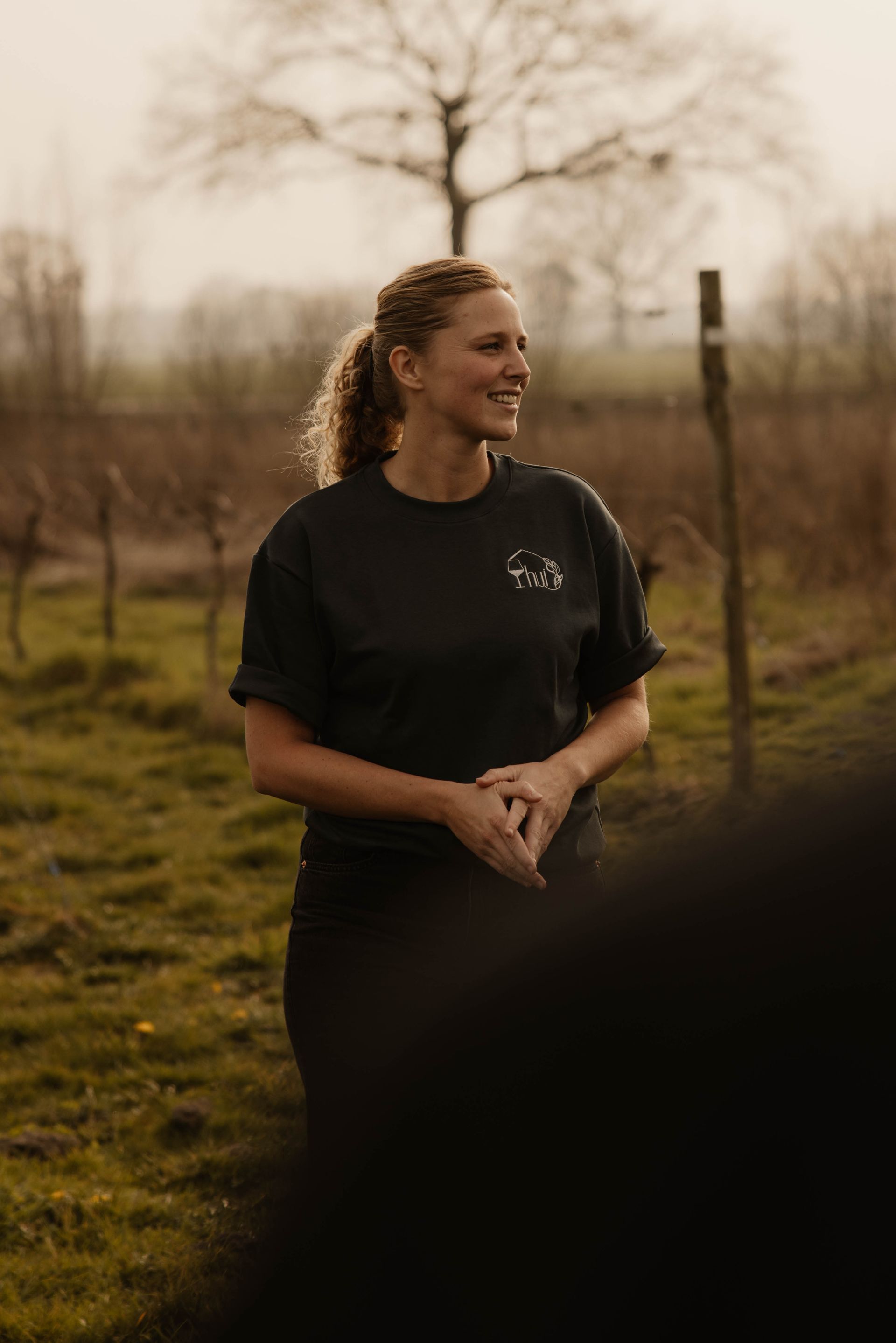 Een persoon staat in een donker shirt buiten, glimlachend in een wijngaard.