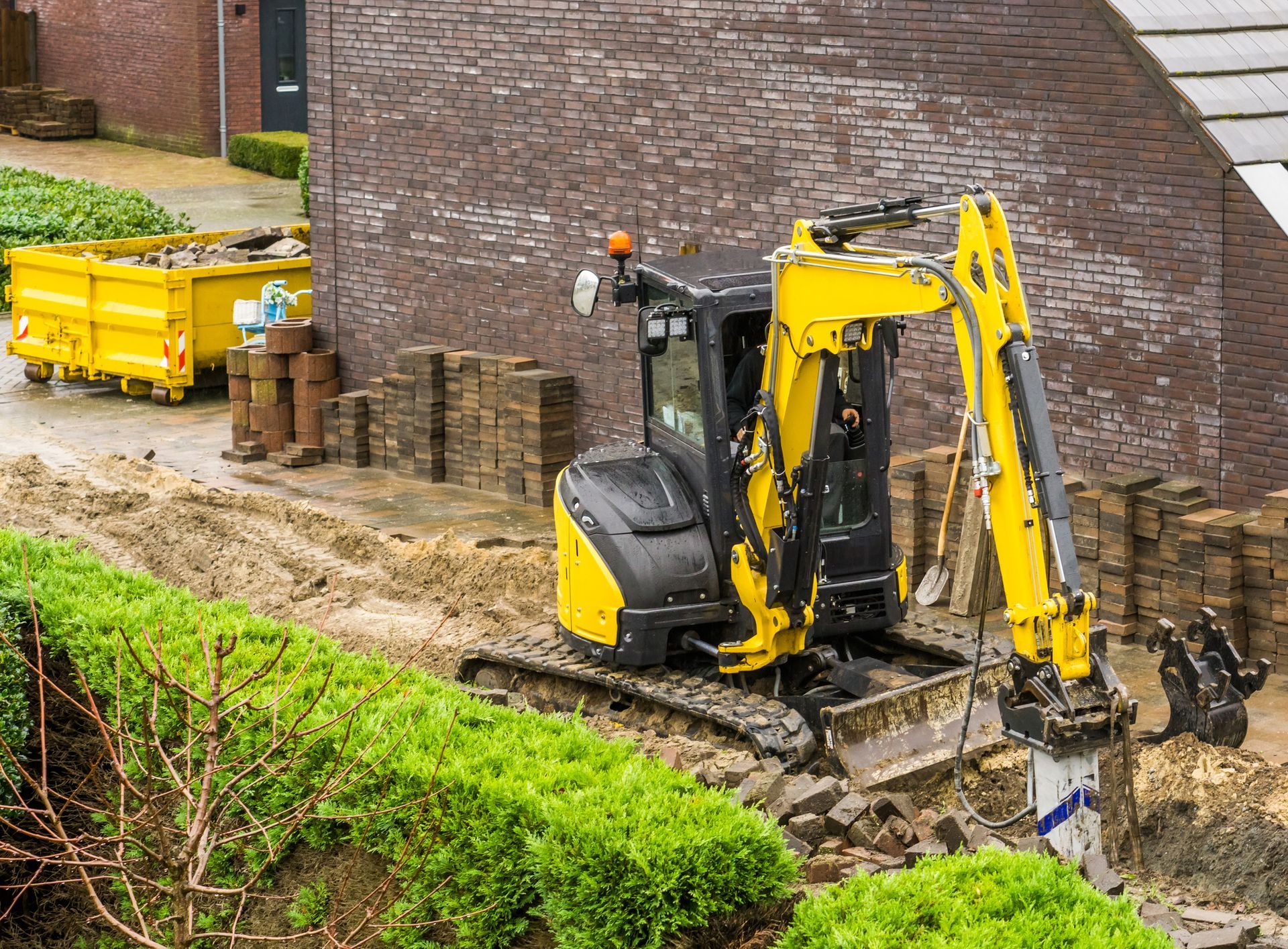 Yellow excavator digging near brick wall, with yellow dumpster in background.