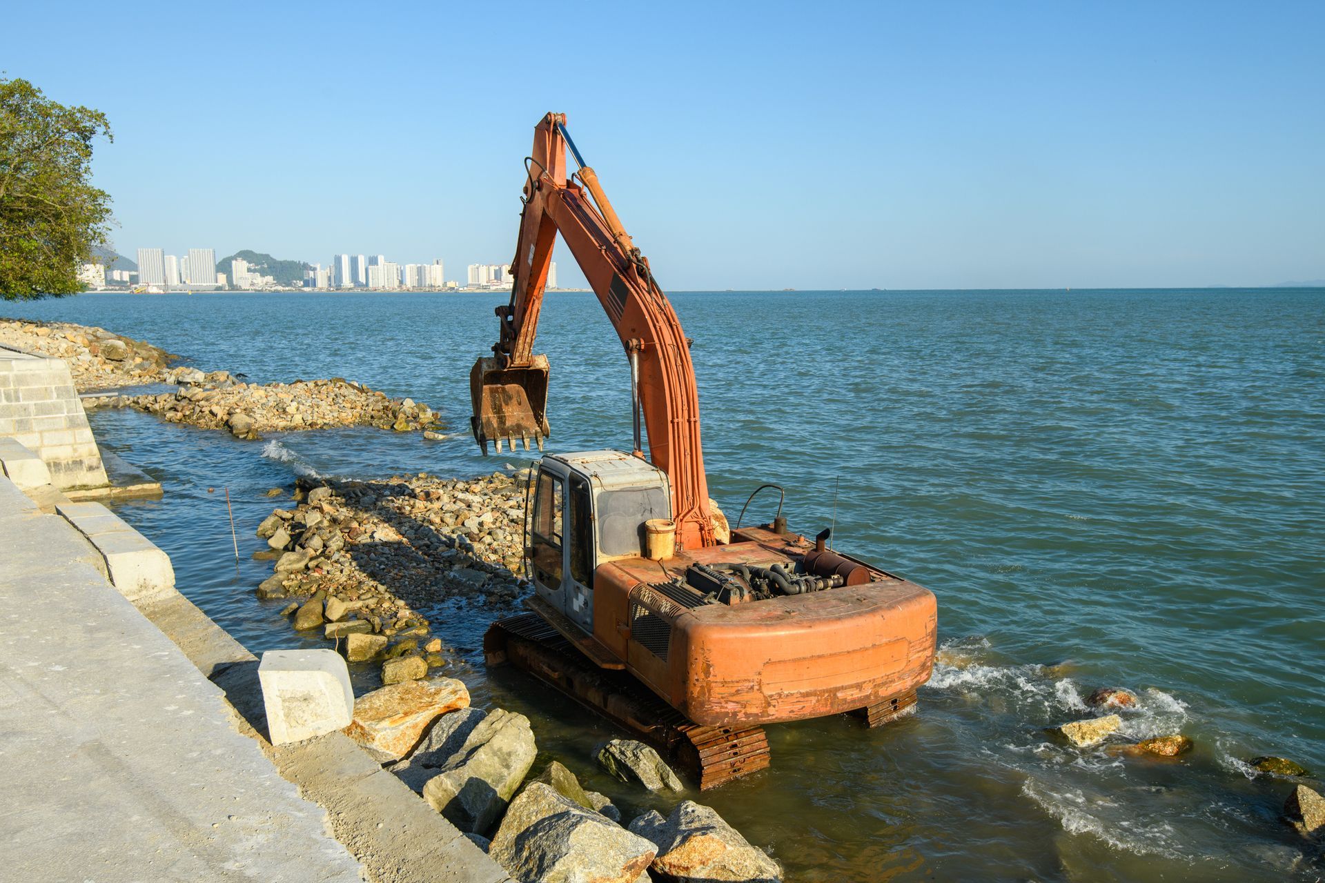 Orange excavator working in ocean water near a rocky shoreline, with city buildings visible in the distance.