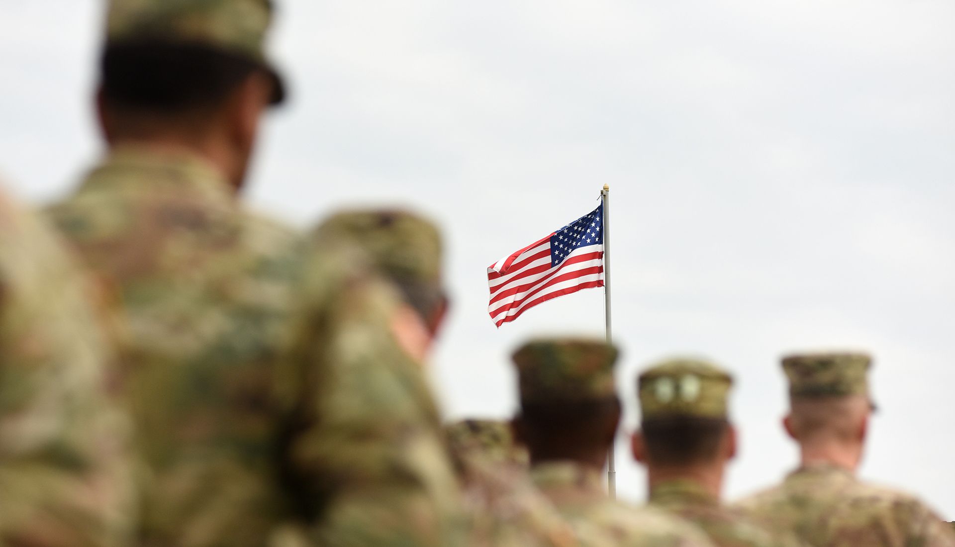 Soldiers in uniform stand at attention, looking at the American flag waving in the sky.