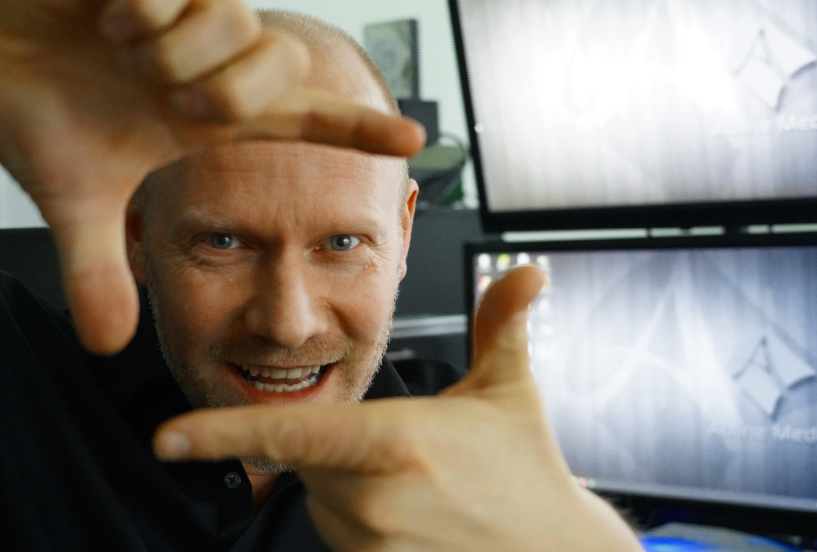 Een lachende man omlijst zijn gezicht met zijn handen in een kantoor, met twee computermonitoren op de achtergrond.
