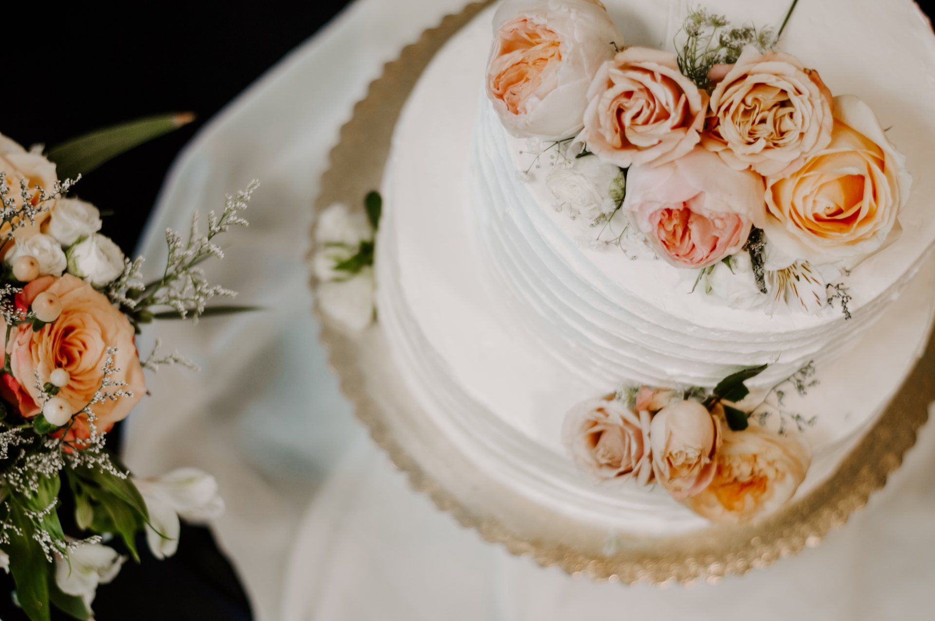 Een witte bruidstaart met perzikkleurige rozen en groen op een tafel, met een bloemenboeket ernaast.