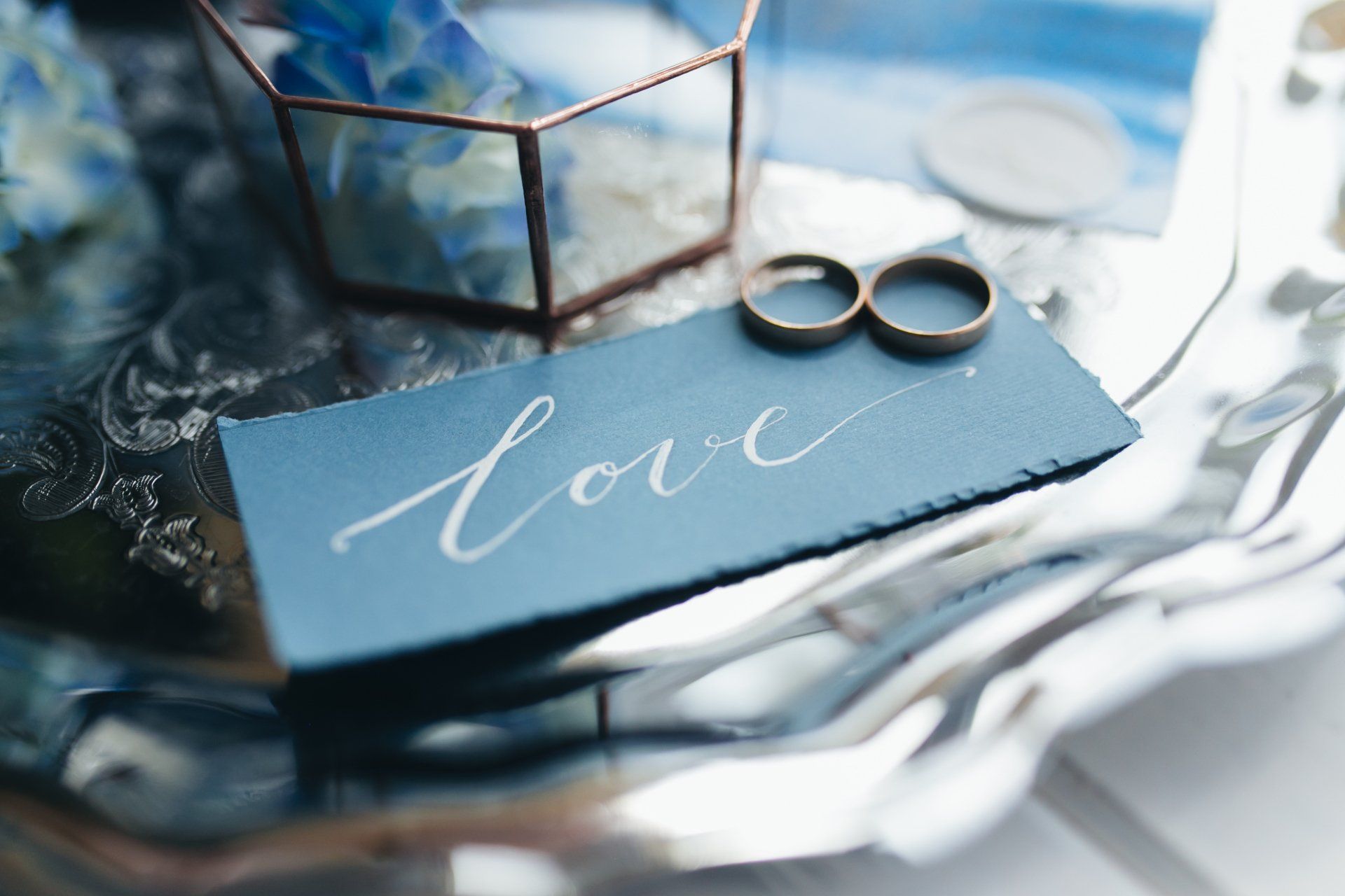 Blauwe kaart met het woord 'liefde' in witte letters naast trouwringen op een reflecterend dienblad.