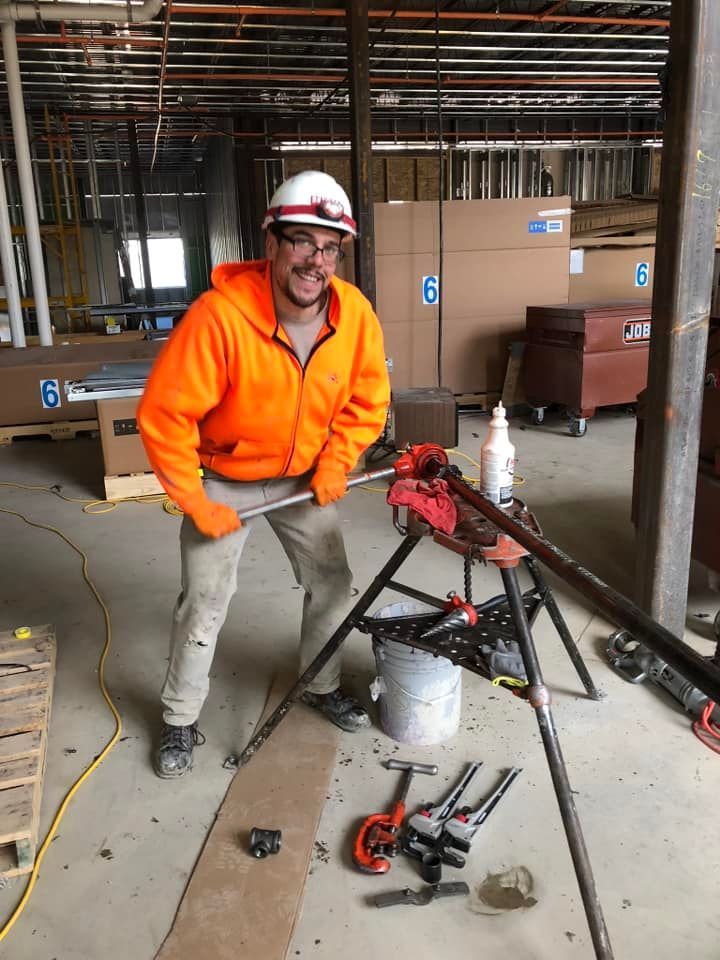 A man in an orange jacket is standing next to a pipe clamp in a room.