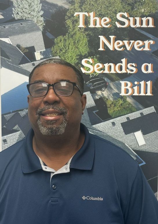 Man in navy polo smiling before a solar panel backdrop with the text “The Sun Never Sends a Bill”