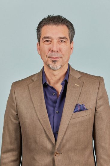 Man in a tan blazer and navy shirt standing against a light gray background