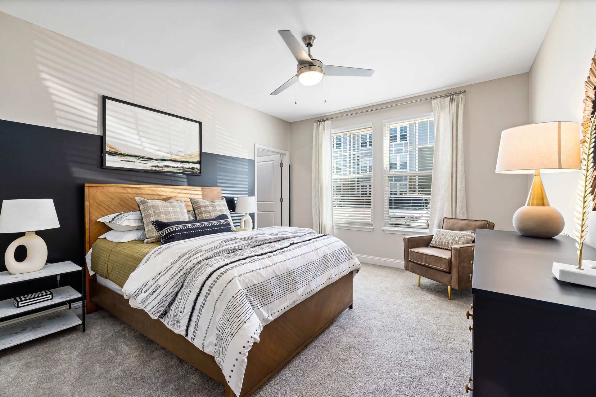 Bright bedroom with a made bed, gray carpet, black dresser, ceiling fan, and window seat.