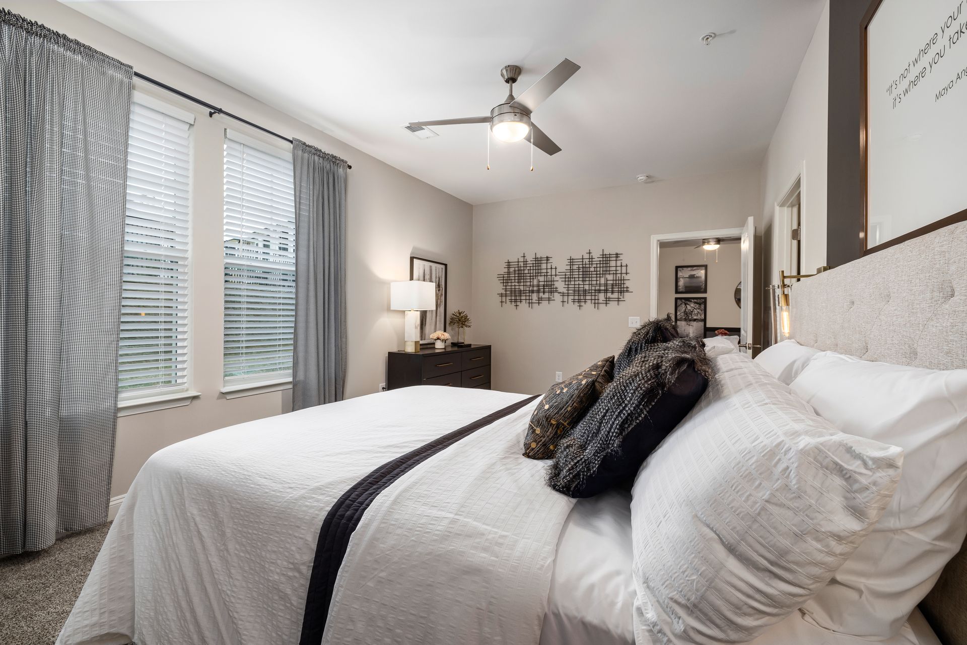 Bright bedroom with a large bed, white bedding, ceiling fan, and window with patterned curtains.