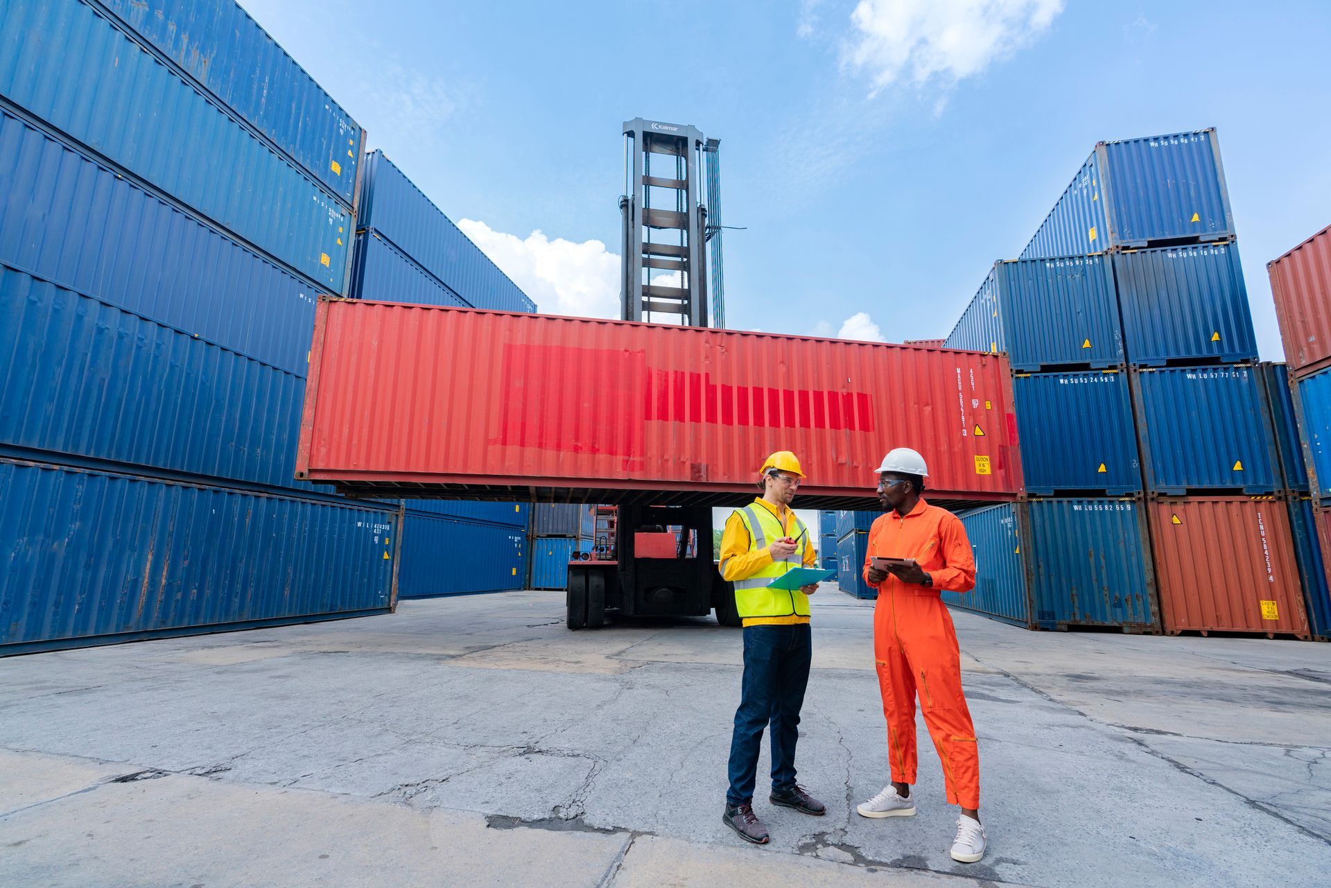 Two workers are seen in front of commercial storage containers at a port. Two workers are seen in front of commercial storage containers at a port.