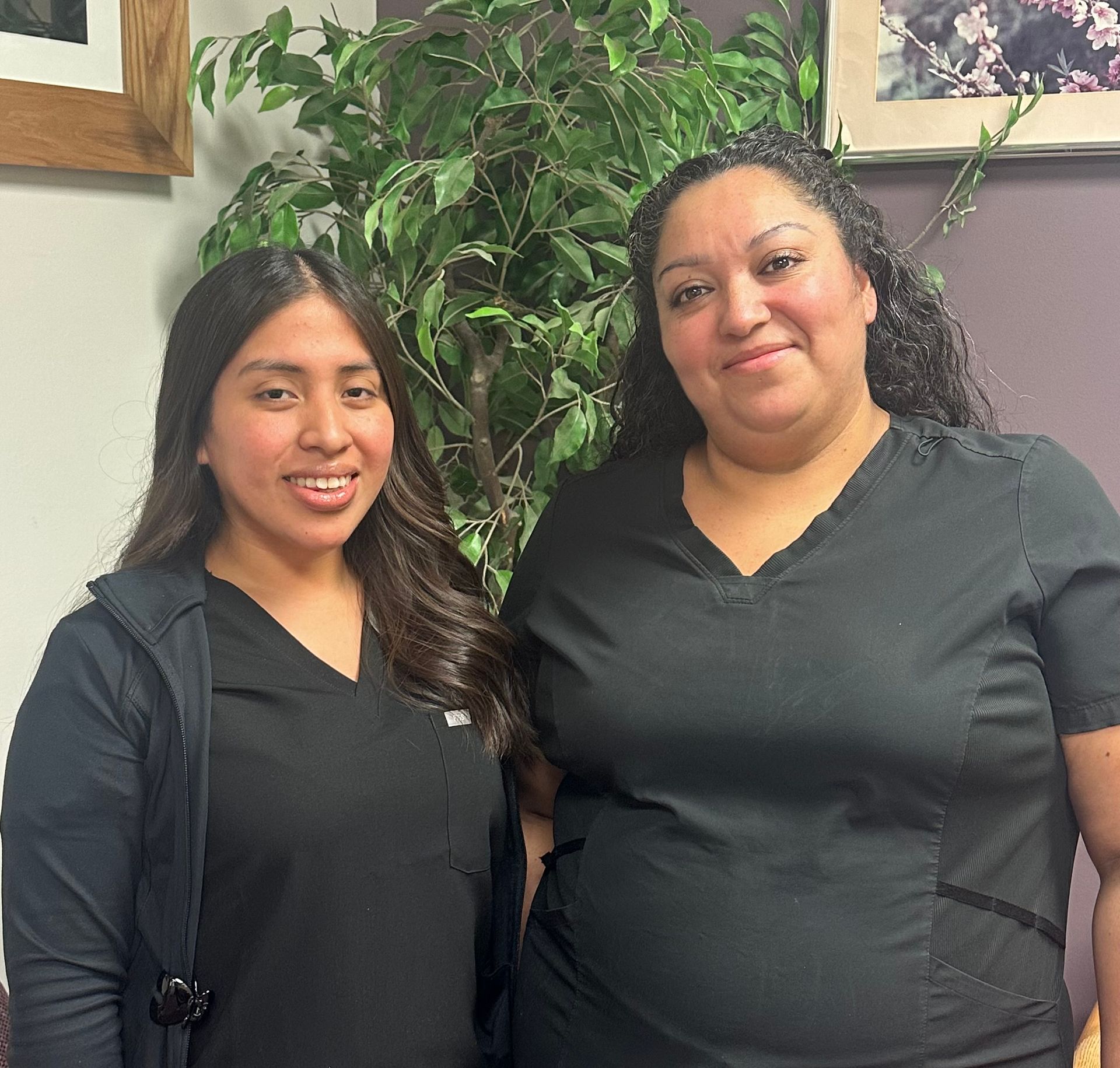Two women in black medical scrubs smiling near a leafy plant, indoor setting.
