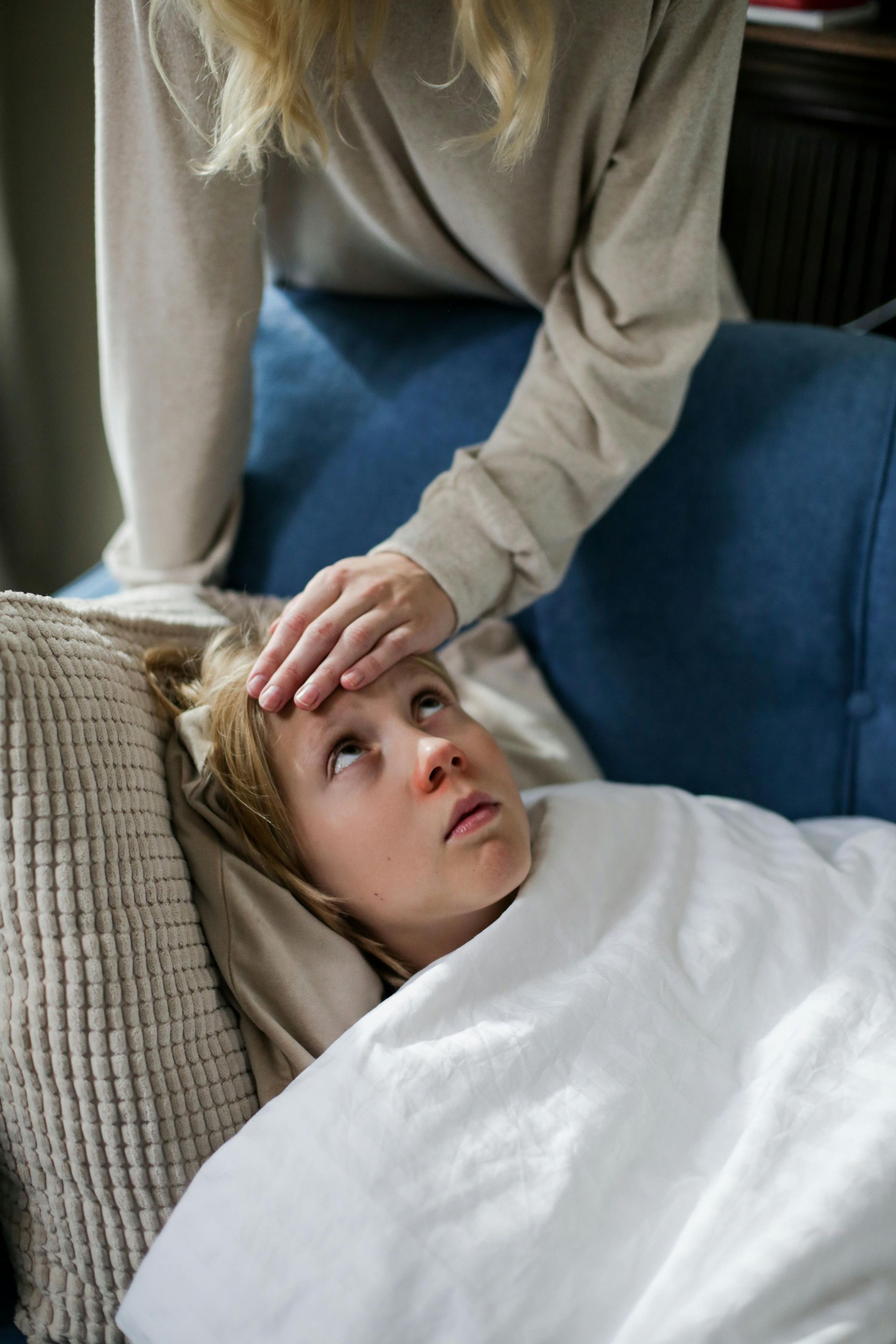 A child is sick on a couch and a mother is placing her hand on the child's forehead.
