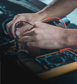 Hands using probes to test an electronic circuit board with a multimeter and red wire leads | E&R Automotive