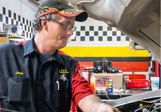 Mechanic in black and red uniform works under a car hood in a garage | E&R Automotive