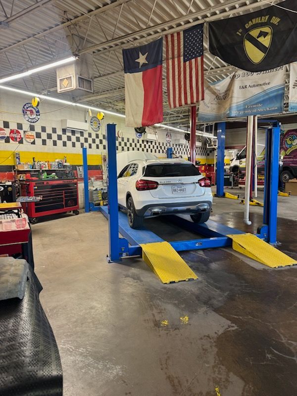 White car on blue lift in auto repair shop with yellow ramps and Texas flag overhead | E&R Automotive