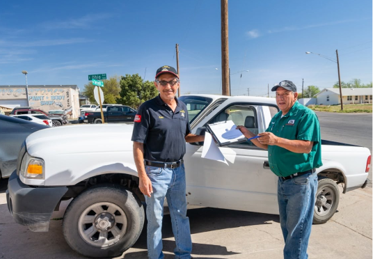 Two men standing beside a white pickup truck in a parking lot, one holding paperwork. | E&R Automotive