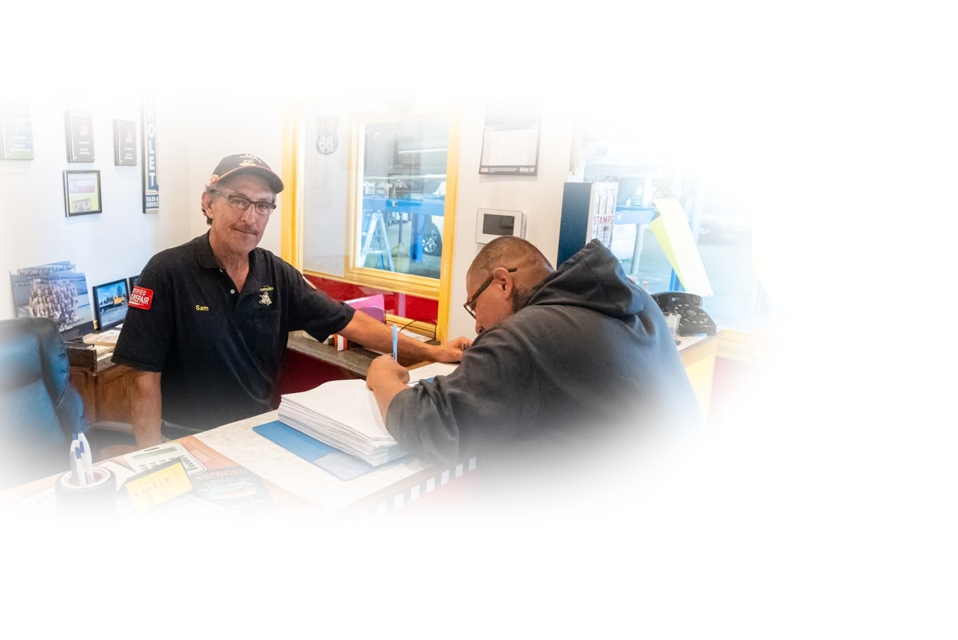 Two people at a service counter exchanging papers in a small office or shop | E&R Automotive