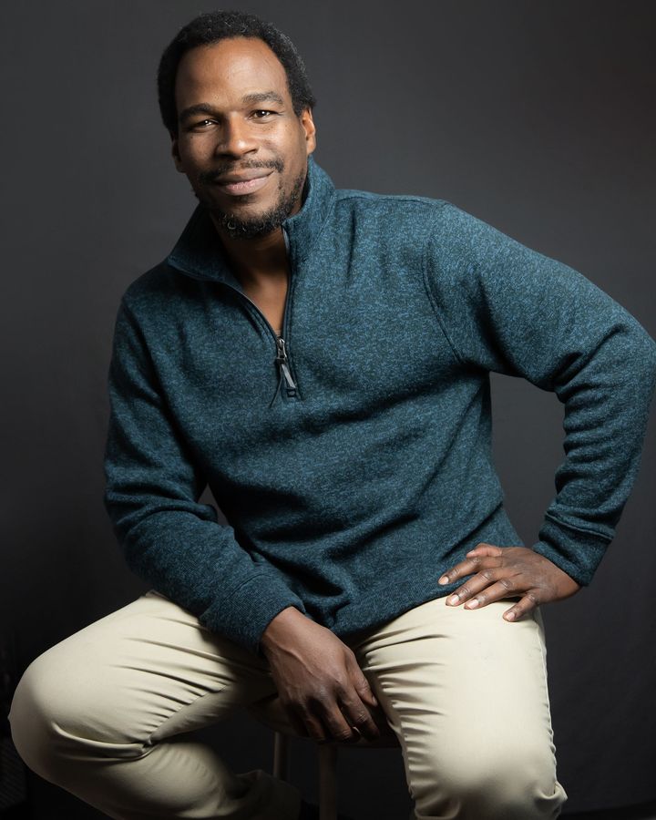 A man in a blue sweater is sitting on a stool with his hands on his hips.