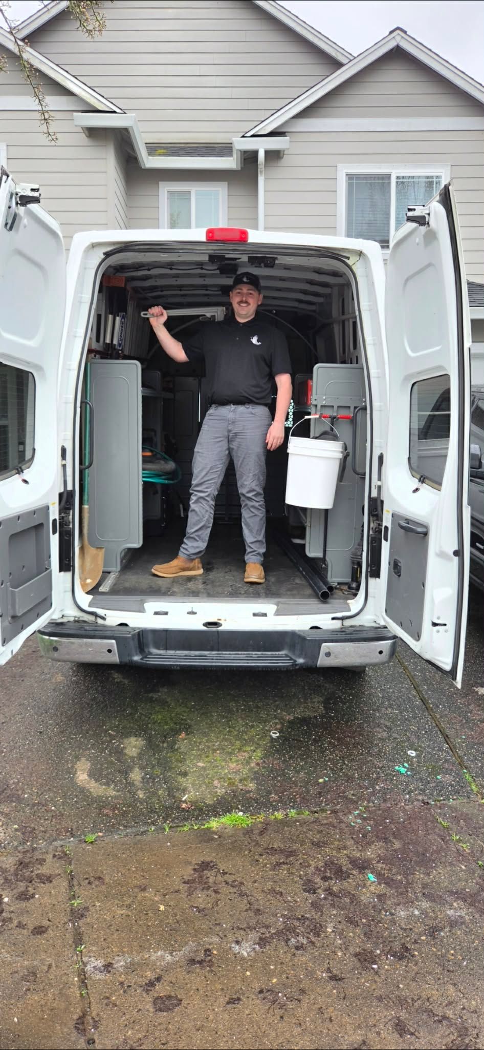 A worker in a black shirt and grey pants stands inside the back of an open white utility van parked in a driveway.