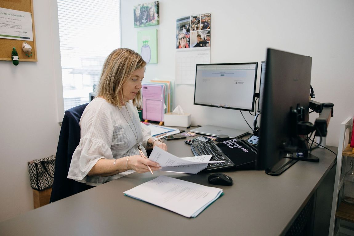 Woman working at a desk with dual monitors in an office, reviewing papers and typing on a keyboard.