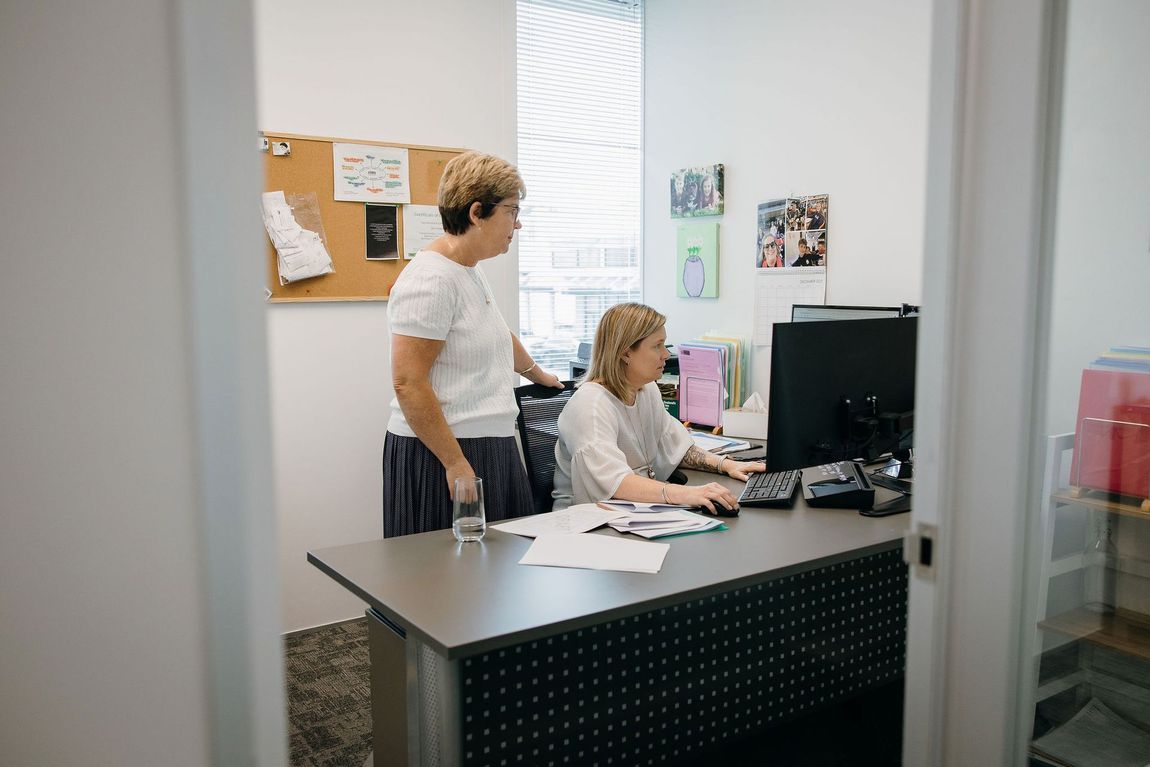 Two coworkers at desks in a small office, one seated at a computer and one standing nearby.