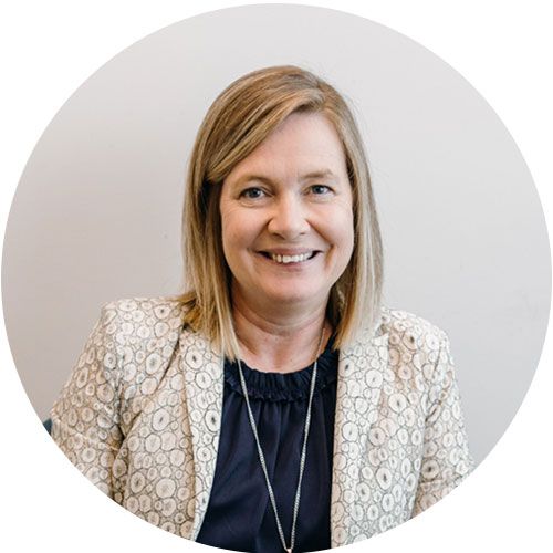 Smiling woman with blonde hair wearing a beige patterned blazer and dark top against a light gray background