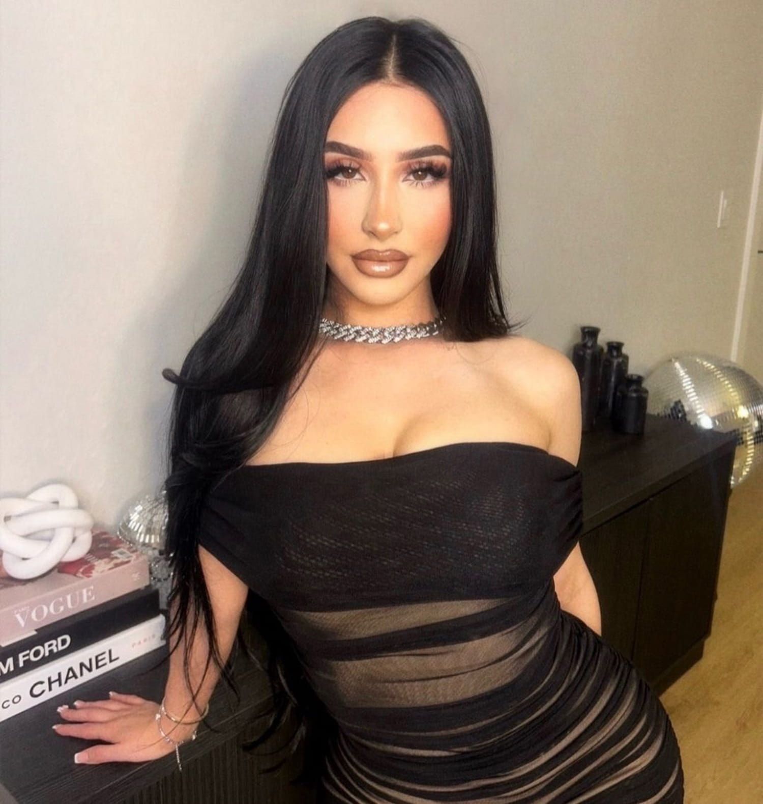 Woman in a black off-shoulder dress and choker, posing indoors beside stacked books.