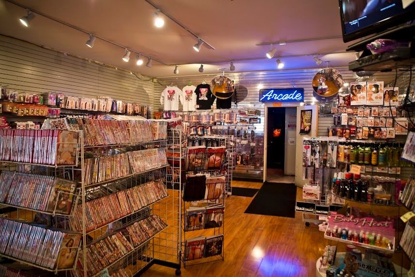 Cluttered gift shop with shelves of souvenirs, warm lights, wooden floor, and a blue neon sign above the back door