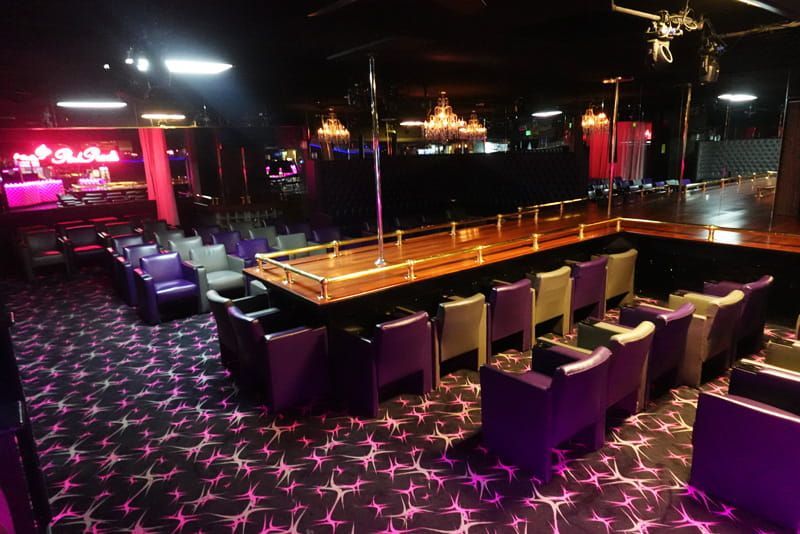 Dim nightclub with a long illuminated bar, purple seating, and pink patterned carpet