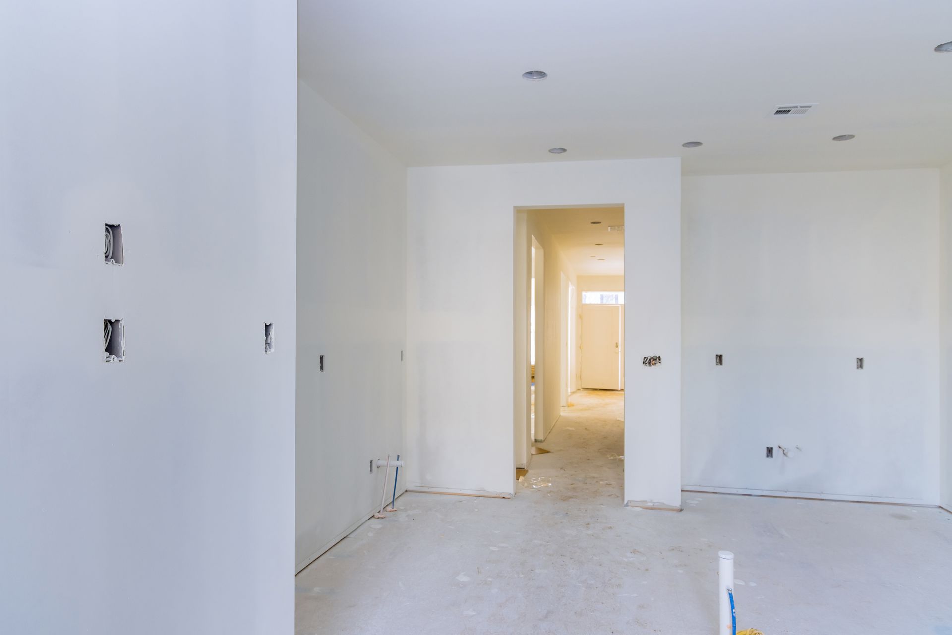 Interno di una stanza in costruzione, pareti dipinte di bianco, con prese elettriche e una porta che conduce a un corridoio.
