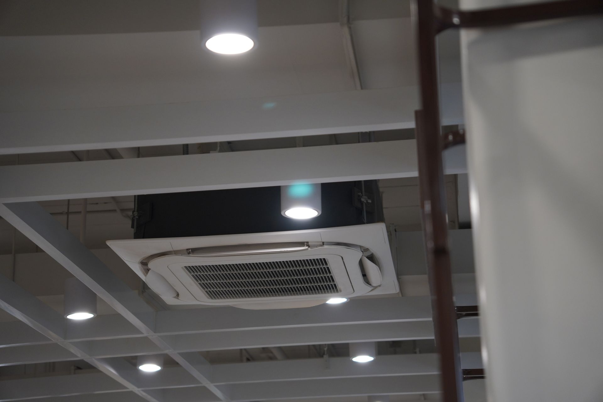 Unità di aria condizionata montata a soffitto. Scatola nera e griglia bianca. Interno con travi, luci e superfici bianco sporco.