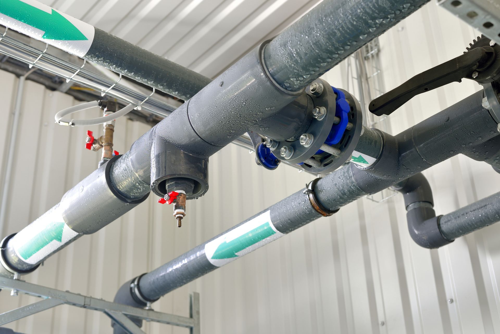 Tubi grigi con valvole e frecce di flusso verdi, montati su una parete industriale bianca.