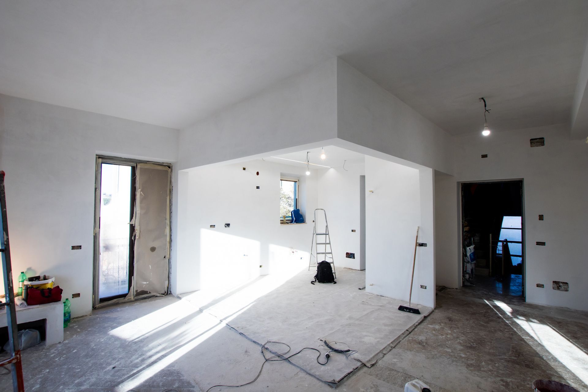 Interno di una stanza in fase di ristrutturazione, con pareti bianche, pavimento nudo e materiali da costruzione.