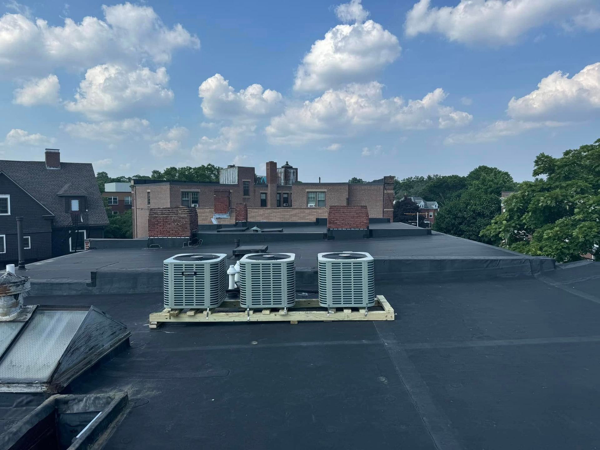 Rooftop with air conditioning units on a wooden pallet; other buildings and blue sky with clouds in the background.