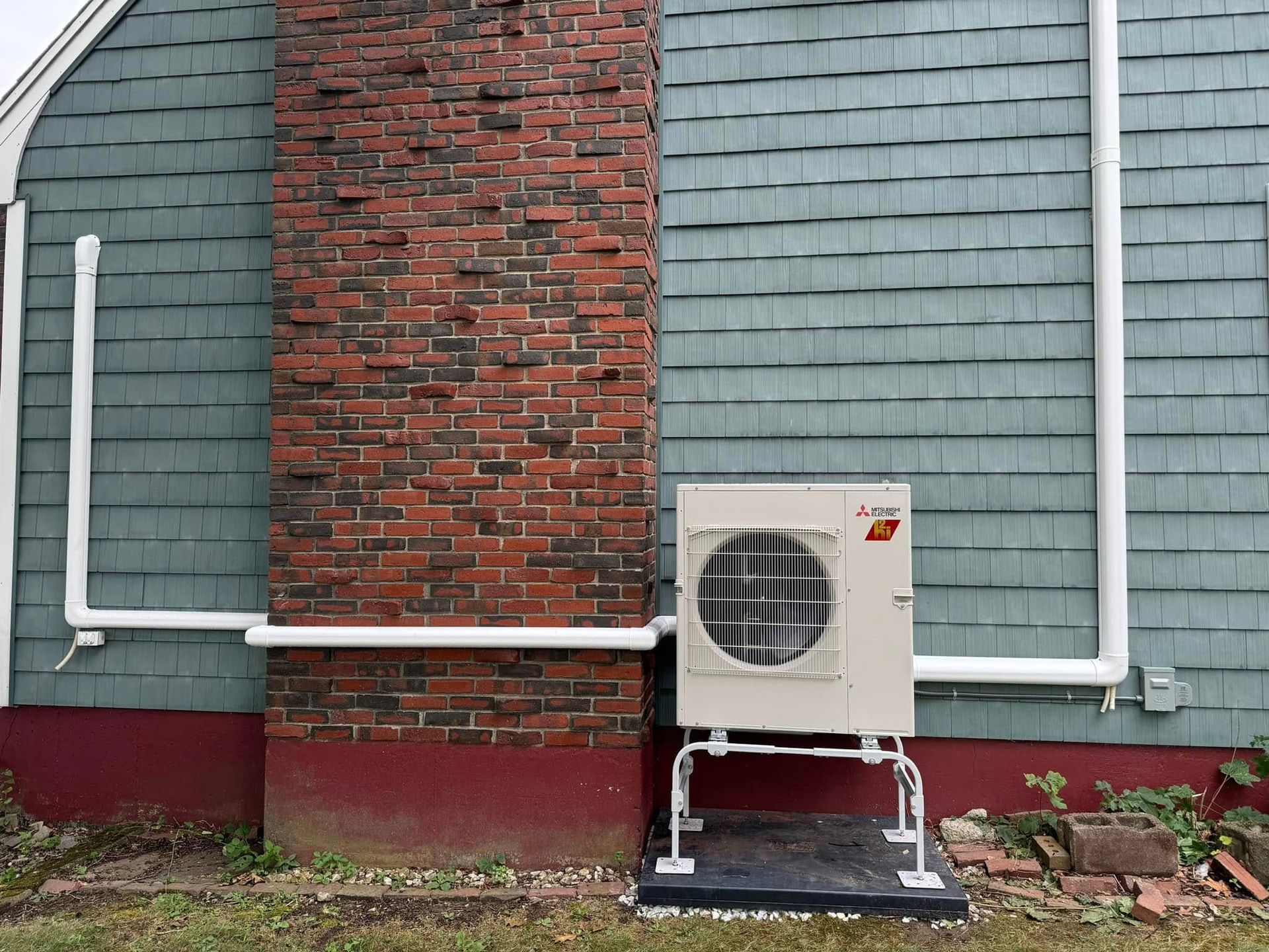 An air conditioning unit on a stand, mounted on a brick chimney and blue siding, with white pipes.