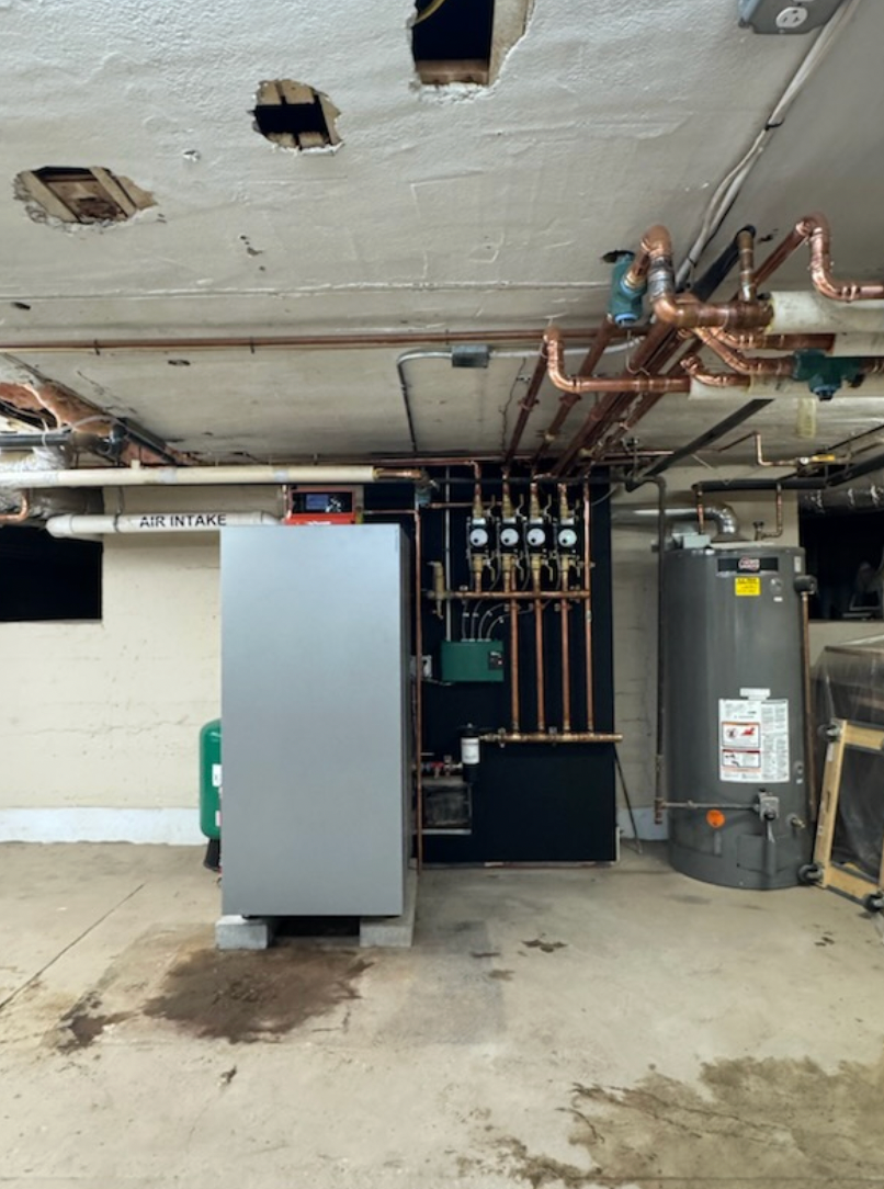 Basement with a large silver tank, pipes, and a water heater. Concrete floor, damaged ceiling.