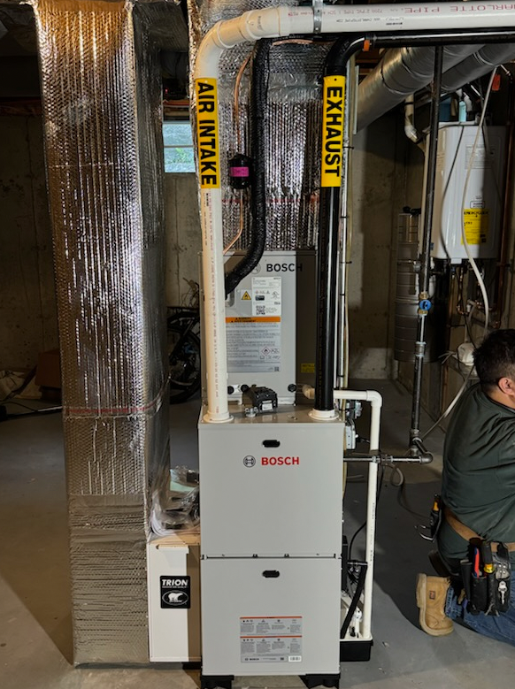 A Bosch furnace with air intake and exhaust pipes. A worker is in the frame. The setting appears to be a basement.