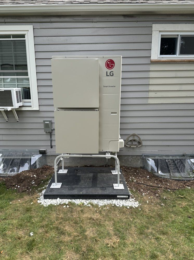 An LG outdoor heat pump unit on a gray house, set on a raised black platform, in a grassy yard.