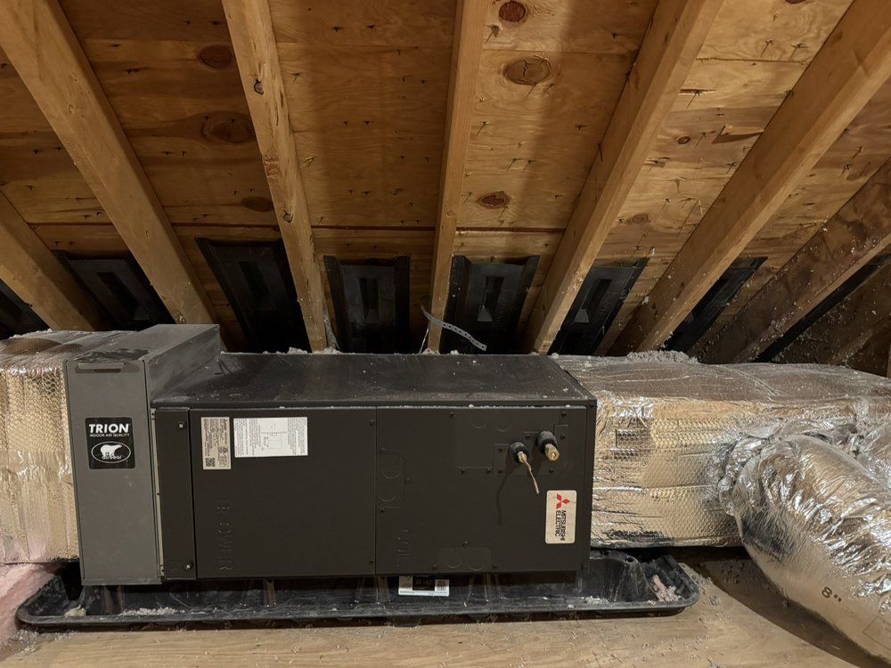 Furnace in attic, rectangular and dark gray, on a black tray. Wood beams overhead.