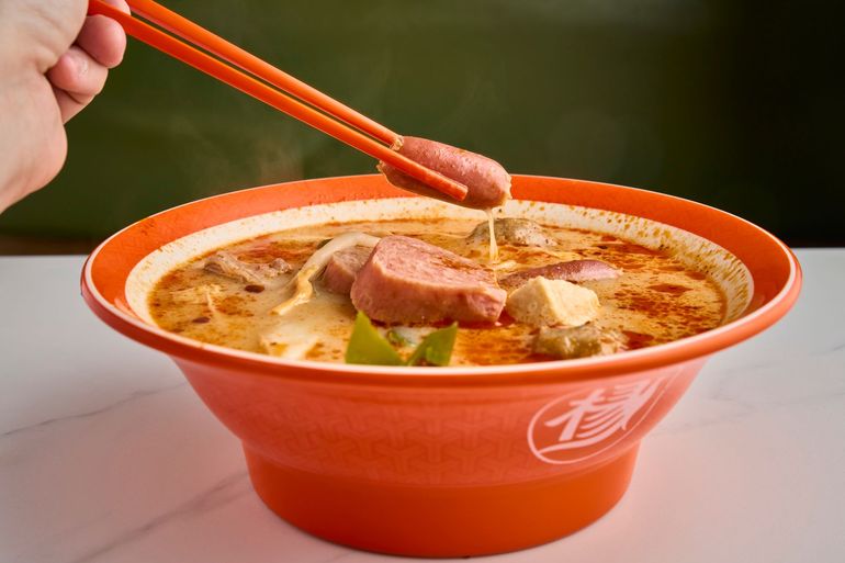 Red bowl of noodle soup with chopsticks lifting sausage over a tabletop