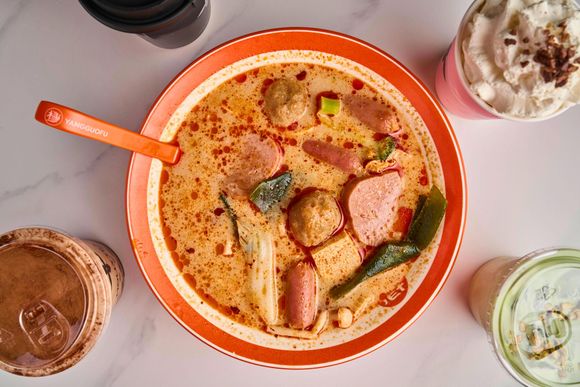 Orange bowl of creamy soup with tofu and vegetables, surrounded by three iced drinks on a table